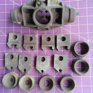 May include: A collection of vintage, rusty metal tools and parts, including a large, central piece with a circular opening and several smaller, rectangular and circular components. The tools are arranged on a purple grid surface.