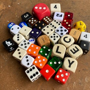May include: A large collection of assorted dice in various colors, sizes, and materials. The pile includes standard numbered dice, translucent red and green dice, wooden dice with letters spelling "CLE" and "FAST", and dice with symbols and text.
