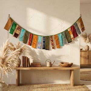 May include: A colorful textile garland hangs on a white wall. The garland features rectangular fabric pieces in various colors and patterns, strung along a green cord. A wooden bench holds books, a watering can, and a wooden bowl.