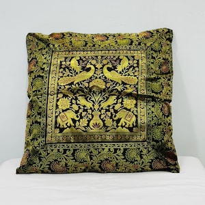 May include: Square cushion with a black background and an ornate gold and green floral and animal pattern. The design includes peacocks, elephants, and floral motifs. The cushion is placed on a white surface.