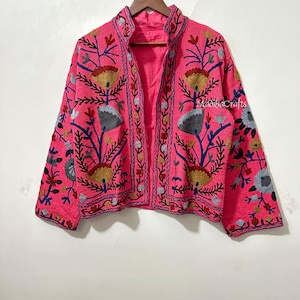 Op de afbeelding: Een felroze jasje met ingewikkelde bloemenborduurwerk in geel, blauw en grijs. De jas heeft een hoge kraag en hangt aan een houten kleerhanger. De borduursels zijn zichtbaar op de mouwen en de voorkant. De merknaam "MadihaCrafts" is zichtbaar.