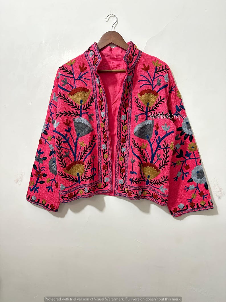 Op de afbeelding: Een felroze jasje met ingewikkelde bloemenborduurwerk in geel, blauw en grijs. De jas heeft een hoge kraag en hangt aan een houten kleerhanger. De borduursels zijn zichtbaar op de mouwen en de voorkant. De merknaam "MadihaCrafts" is zichtbaar.