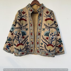 Op de afbeelding: Beige geborduurde jas met een opstaande kraag en bloemenmotief. De jas heeft een beige voering en is versierd met kleurrijk borduurwerk in blauw, rood en groen. De jas hangt aan een houten kleerhanger.