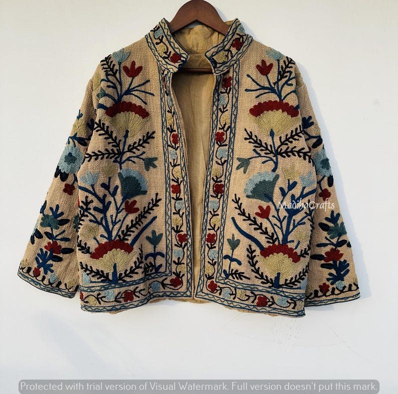 Op de afbeelding: Beige geborduurde jas met een opstaande kraag en bloemenmotief. De jas heeft een beige voering en is versierd met kleurrijk borduurwerk in blauw, rood en groen. De jas hangt aan een houten kleerhanger.