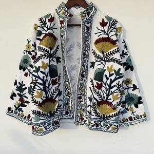 Op de afbeelding: Witte jas met kleurrijke bloemenborduurwerk. De jas heeft een mandarijnkraag en een open voorkant. Het borduurwerk omvat tinten geel, groen, rood en blauw. De jas hangt aan een houten kleerhanger.