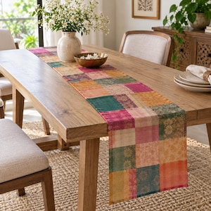 May include: A rectangular table runner featuring a patchwork design of various square fabric panels in shades of pink, orange, teal, and gold. The fabric displays intricate floral patterns and textures, draped across a wooden dining table.