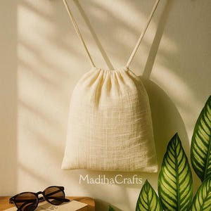 May include: A cream-colored drawstring bag made of natural fabric, hanging from a wooden hanger. The bag is on a wooden table with a pair of sunglasses and a notebook.