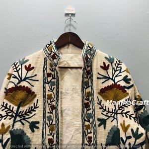 Op de afbeelding: Cr&egrave;mekleurige geborduurde jas met een hoge kraag. De jas heeft een bloemenmotief in tinten geel, bordeaux, blauw en groen. De jas hangt aan een houten hanger.