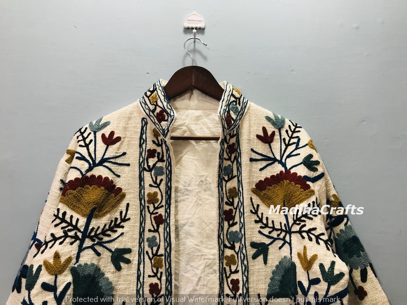 Op de afbeelding: Cr&egrave;mekleurige geborduurde jas met een hoge kraag. De jas heeft een bloemenmotief in tinten geel, bordeaux, blauw en groen. De jas hangt aan een houten hanger.