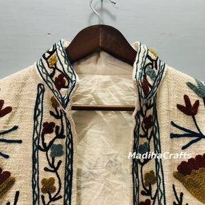 Op de afbeelding: Cr&egrave;mekleurige geborduurde jas met bloempatronen in blauw, bruin en geel. De jas hangt aan een houten kleerhanger. De tekst "MadihaCrafts" is zichtbaar op de jas.