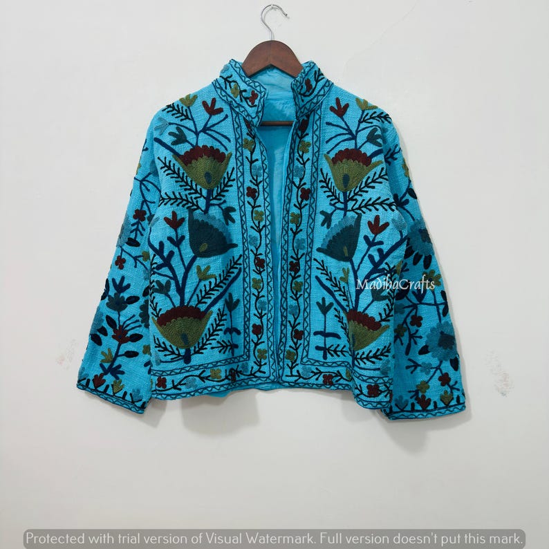 Op de afbeelding: Een lichtblauwe jas met ingewikkelde bloemenborduurwerk in groen, bruin en blauw. De jas heeft een hoge kraag en lange mouwen. De tekst "MadihaCrafts" is zichtbaar op de jas.