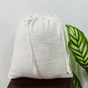 Puede incluir: Una bolsa de cordón blanco hecha de tela texturizada. La bolsa está vacía y tiene un diseño simple.