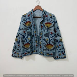 Op de afbeelding: Lichtblauwe jas met bloemenborduurwerk in geel, rood en blauw. De jas heeft een opstaande kraag en is aan de voorkant open. De tekst "MadihaCrafts" is zichtbaar. Hij hangt aan een houten hanger.