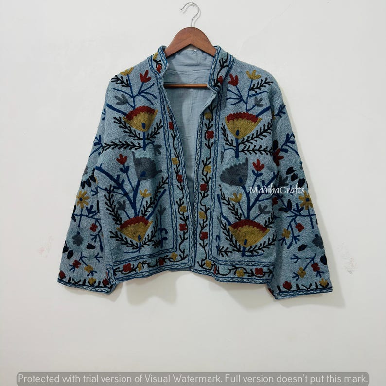 Op de afbeelding: Lichtblauwe jas met bloemenborduurwerk in geel, rood en blauw. De jas heeft een opstaande kraag en is aan de voorkant open. De tekst "MadihaCrafts" is zichtbaar. Hij hangt aan een houten hanger.