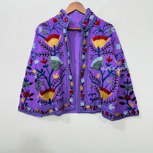 Op de afbeelding: Een levendige paarse jas met ingewikkelde bloemenborduursels in geel, rood en blauw. De jas heeft een opstaande kraag en wordt op een houten hanger getoond. De tekst "MadihaCrafts" is zichtbaar.
