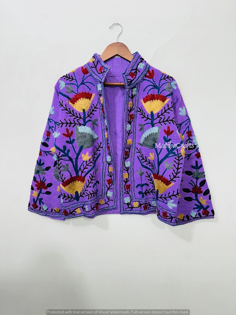 Op de afbeelding: Een levendige paarse jas met ingewikkelde bloemenborduursels in geel, rood en blauw. De jas heeft een opstaande kraag en wordt op een houten hanger getoond. De tekst "MadihaCrafts" is zichtbaar.