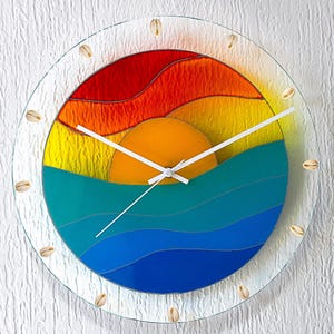 May include: A circular wall clock made of fused glass featuring a vibrant sunset and ocean scene with layered bands of red, orange, yellow, teal, and blue. The clear glass border is decorated with small, light-colored seashells and white hands.