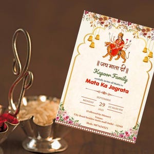 May include: An invitation card with floral and gold accents, featuring an illustration of a deity riding a tiger. The card includes text in Hindi and English, with the date and time of an event. Two silver bowls with red powder and rice are in the foreground.