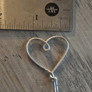 Puede incluir: Un amuleto de coraz&oacute;n de alambre plateado con un bucle en la parte superior y una cola de alambre larga y delgada.