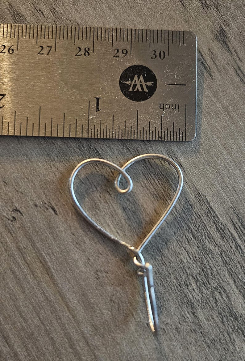 Puede incluir: Un amuleto de coraz&oacute;n de alambre plateado con un bucle en la parte superior y una cola de alambre larga y delgada.