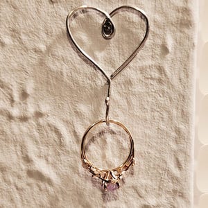 Puede incluir: Un soporte para anillos de alambre en forma de coraz&oacute;n plateado con una peque&ntilde;a cuenta negra en la parte superior. Un anillo de oro con una piedra morada y peque&ntilde;as piedras transparentes cuelga del soporte.