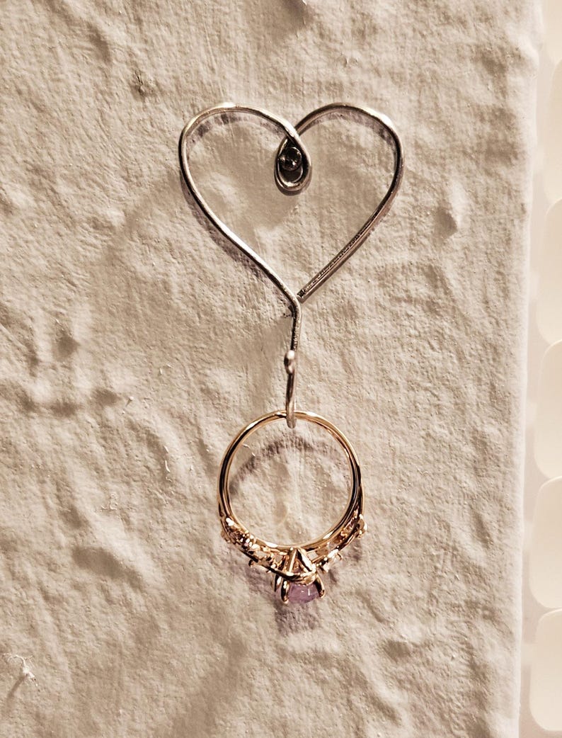 Puede incluir: Un soporte para anillos de alambre en forma de coraz&oacute;n plateado con una peque&ntilde;a cuenta negra en la parte superior. Un anillo de oro con una piedra morada y peque&ntilde;as piedras transparentes cuelga del soporte.