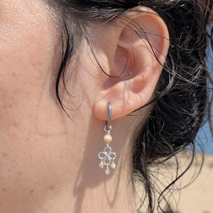 Peut inclure: Une boucle d'oreille créole argentée avec une breloque pendante composée d'une petite perle ronde couleur crème en haut, d'un nœud central en fil métallique et de trois minuscules perles rondes couleur crème suspendues en bas.