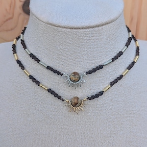 Peut inclure: Un collier à deux rangs avec des cordons noirs et des accents dorés et argentés. Chaque cordon a un pendentif en forme de soleil avec une pierre brune au centre.