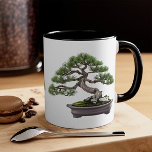 Könnte beinhalten: Eine weiße Keramiktasse mit schwarzem Rand und Henkel zeigt eine farbenfrohe Illustration eines Bonsai-Baums in einem grauen Topf. Die Tasse steht auf einer Holzoberfläche.