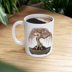 May include: White ceramic mug with a black handle featuring a bonsai tree with weeping branches in a brown pot. The mug is filled with black coffee.