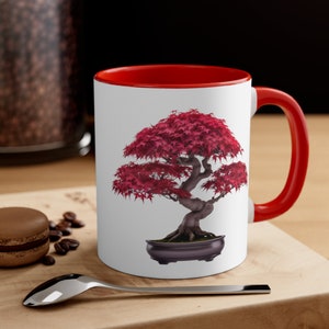 Puede incluir: Taza de cerámica blanca con borde y asa rojos. La taza presenta una imagen colorida de un bonsái con hojas rojas en una maceta negra.