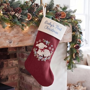 May include: A deep red Christmas stocking featuring a white cuff embroidered with the text Baby's First Christmas in blue. The body of the stocking displays an embroidered white lamb resting within a circular wreath of blue and green flowers.