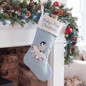 May include: A light blue Christmas stocking with a white cuff embroidered with the text Baby's First Christmas. The stocking features an embroidered penguin on a snowy hill, accented with small white buttons, silver snowflakes, and red berries.