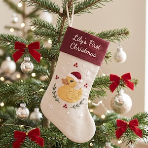 Puede incluir: Un calcetín navideño de color crema con un puño de terciopelo rojo oscuro bordado con el texto Lily's First Christmas. El cuerpo del calcetín muestra un patito de goma amarillo bordado con un gorro de Papá Noel rojo, rodeado de hojas verdes, bayas rojas y copos de nieve blancos.
