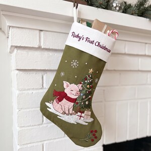 May include: An olive green Christmas stocking with a white cuff embroidered with the text Ruby's First Christmas. The stocking features an embroidered pink pig wearing a red scarf, a decorated Christmas tree, snowflakes, and gift boxes.