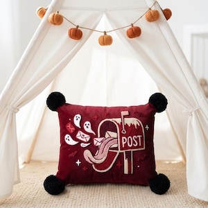May include: A rectangular dark red throw pillow with black pom-poms at each corner. The front features an embroidered design of a mailbox with the word POST, surrounded by white ghosts, envelopes, and small red stars on a textured fabric surface.