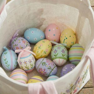 May include: A collection of painted Easter eggs in a white fabric basket. The eggs are decorated with floral patterns, stripes, and polka dots in pastel colors like pink, blue, yellow, and lavender. Some eggs have ribbons.