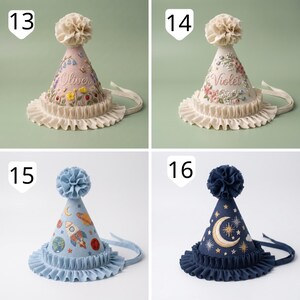 May include: Four cone-shaped party hats with ruffled brims and pom-pom toppers. Hat 13 is pink with floral embroidery and the name Oliver. Hat 14 is cream with floral embroidery and the name Violet. Hat 15 is light blue with space motifs. Hat 16 is navy blue with moon and star embroidery.
