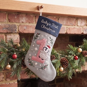 May include: A grey Christmas stocking with a dark blue cuff featuring the embroidered text "Baby's First Christmas". The stocking displays a large pink number one and an embroidered penguin wearing a red hat, surrounded by small white snowflakes.