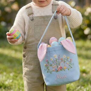 May include: A light blue Easter basket with pink bunny ears and a floral embroidery design. The basket is filled with colorful Easter eggs. The name "Mia" is embroidered on the front. An Easter egg is held in the other hand.