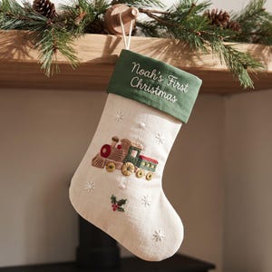 Puede incluir: Un calcetín navideño de color crema con un puño verde bordado con el texto Noah's First Christmas. El cuerpo del calcetín muestra un tren de vapor bordado en rojo, verde y marrón, acentuado con copos de nieve blancos y hojas de acebo.
