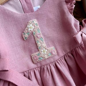 May include: A dusty rose-colored dress with a floral embroidered number "1" on the front. The dress features ruffled sleeves and a matching sash. The embroidered number is detailed with white, pink, and green floral accents, creating a delicate and charming design.