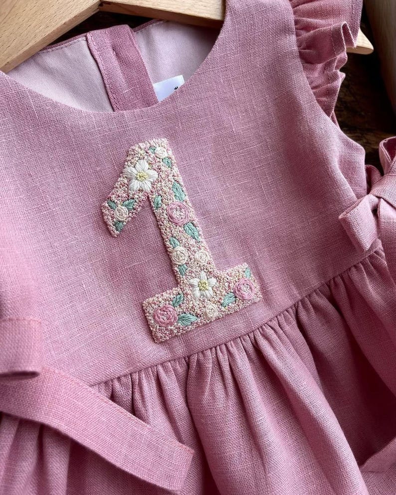May include: A dusty rose-colored dress with a floral embroidered number "1" on the front. The dress features ruffled sleeves and a matching sash. The embroidered number is detailed with white, pink, and green floral accents, creating a delicate and charming design.