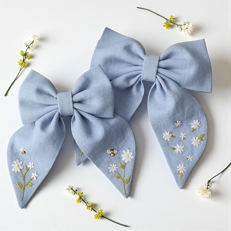 May include: Two light blue linen hair bows with embroidered white daisies and yellow bees. The bows have long, flowing tails. Sprigs of yellow and white flowers are scattered around the bows on a white background.