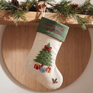 May include: A cream-coloured fabric Christmas stocking featuring a green velvet cuff embroidered with the text Anna's First Christmas. The stocking displays an embroidered green pine tree with a red bow, three small gift boxes, and snowflakes.