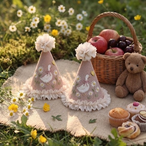 May include: Two cone-shaped party hats in dusty pink felt, each topped with a cream pom-pom and a ruffled trim. The hats feature embroidered white geese, yellow butterflies, and daisy flowers. The hats are displayed on a hessian mat.