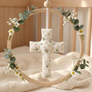 Op de afbeelding: Een wit, kruisvormig baby mobile met geborduurde bloemendetails, opgehangen in een houten hoepel versierd met eucalyptus en kleine witte bloemen. De mobiel hangt aan een houten kraal.