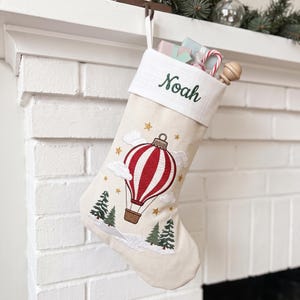 May include: A cream-coloured Christmas stocking featuring an embroidered red and white striped hot air balloon, green pine trees, and gold stars. The white cuff is embroidered with the name Noah in green script and has a subtle plaid pattern.