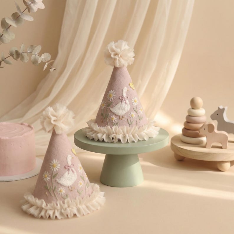 May include: Two cone-shaped party hats in a soft mauve colour, each featuring embroidered white geese, yellow flowers, and green stems. The hats are topped with a ruffled fabric pom-pom and finished with a gathered cream-coloured fabric trim at the base.