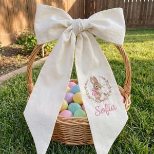 May include: An Easter basket made of woven wicker, filled with pastel eggs, and decorated with a large cream-coloured bow. A cream fabric streamer with a bunny embroidery and the name "Sofia" in pink is attached.